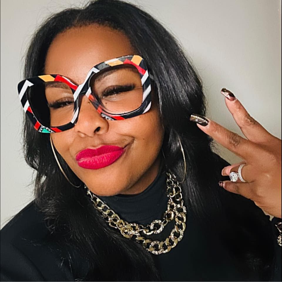 Black woman with colorful patterned glasses, red lipstick, and gold chains, making a peace sign.