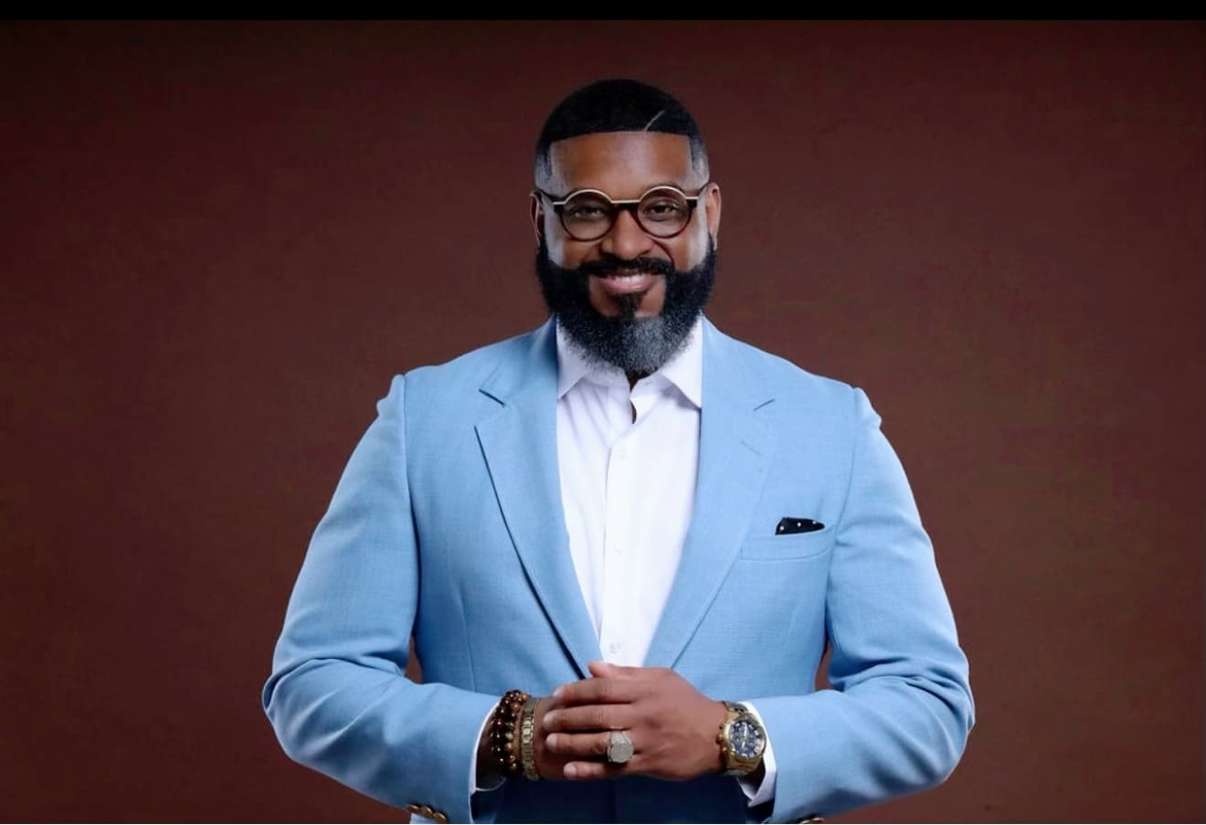 Smiling Black man with a groomed beard and glasses wearing a light blue suit.