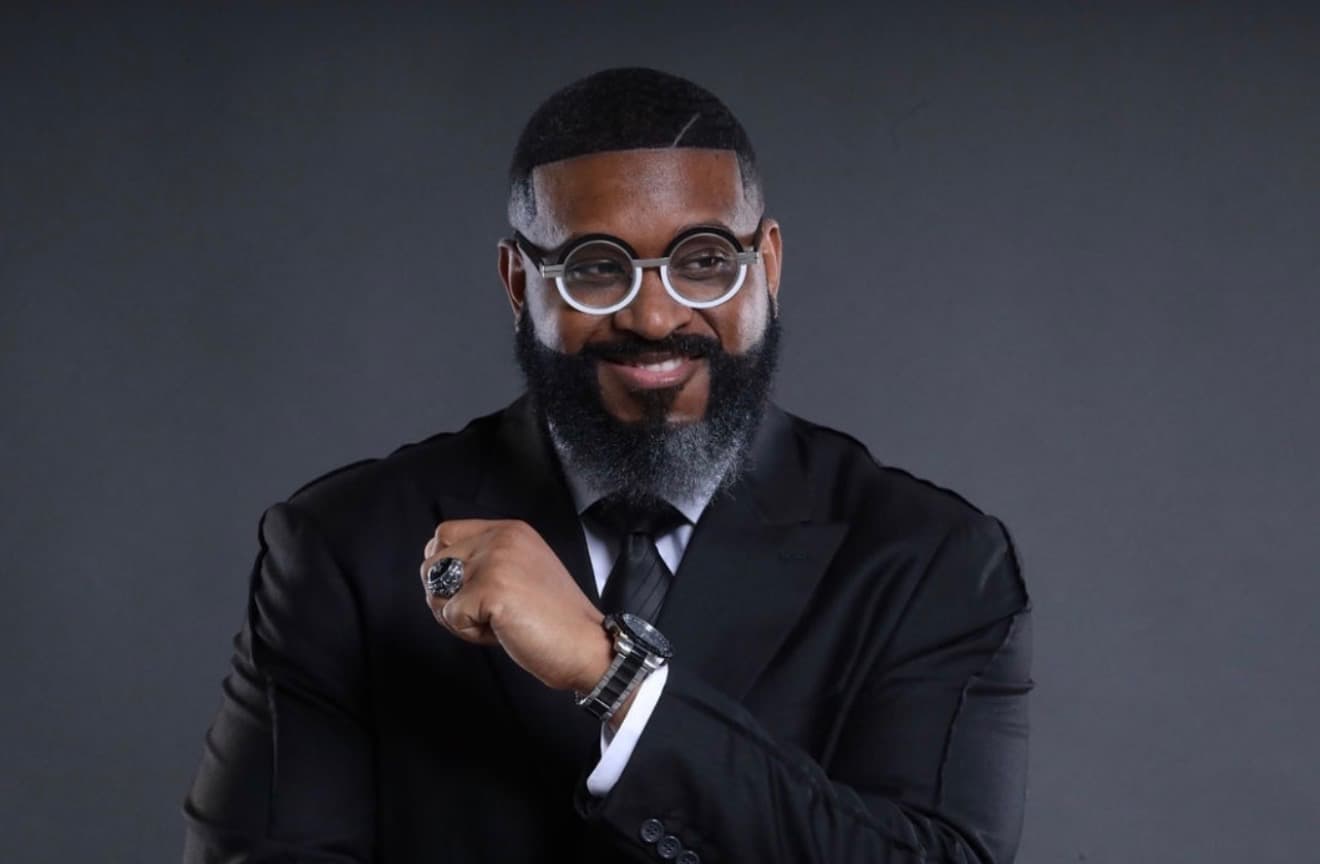 Bearded man in a black suit and round glasses, smiling and showing his jewelry.